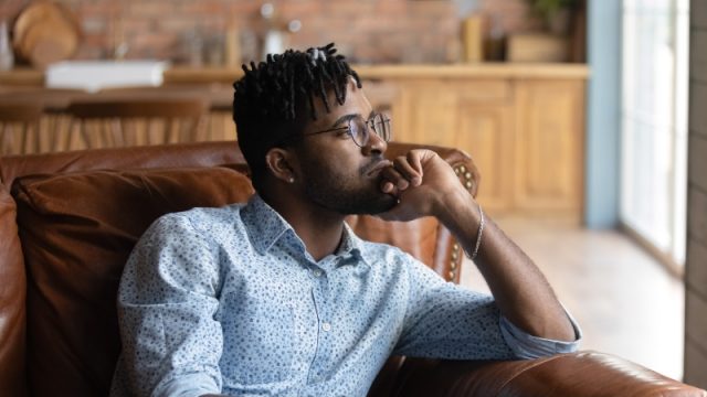 Thoughtful bored millennial guy lost in thoughts, sitting on coach, looking away.