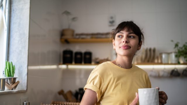 Contemplative young woman drinking coffee or tea and looking window at home